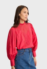 Blusa de algodón roja con un alto cuello rizado, intrincado bordado en la parte delantera y detalles con pliegues. Manga larga abullonada y corte relajado.