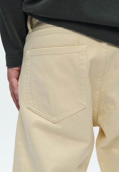 Béžové bavlnené nohavice s vreckom v tvare diamantu na zadnej strane a viditeľným šitím, kombinované s textúrovaným sivým tričkom s dlhým rukávom.