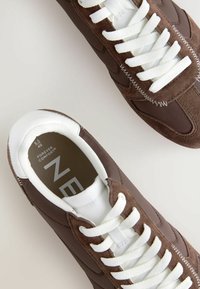 Zapatillas de ante marrón y tela con cordones blancos, que presentan un diseño aerodinámico y costuras en contraste. La etiqueta interior dice "COMODIDAD PARA SIEMPRE."
