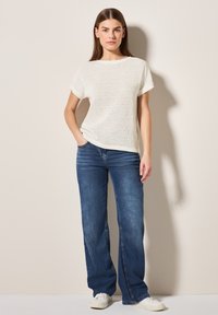 Lätt, stickad vit topp med korta ärmar, kombinerad med blå vida jeans och vita sneakers. Enkel, avslappnad design.