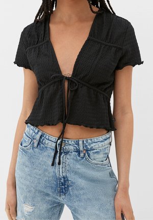 Kobieta nosząca teksturowaną, czarną, krótką bluzkę z krótkim rękawem, zawiązaną z przodu, oraz jasnoniebieskie, przetarte dżinsy z wysokim stanem.