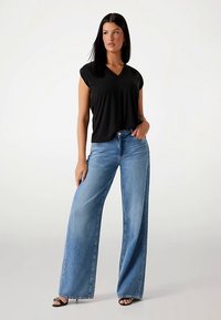Black v-neck top, loose fit; high-waisted light blue wide-leg jeans. Model wearing black strappy sandals, standing against a neutral background.