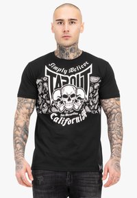 Fekete pamut póló fehér koponya grafikákkal, "TAPOUT" felirattal és bandana-stílusú díszítésekkel. Rövid ujjú és kerek nyakú dizájn.