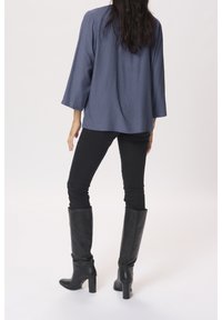 Femme portant un haut à manches longues bleu, un pantalon skinny noir et des bottes noires à talons montantes jusqu'aux genoux, debout de dos face à la caméra.