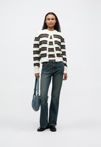 Cardigan a righe bianco e marrone, abbinato a jeans blu e scarpe nere. Il modello tiene in mano una piccola borsa blu. Sfondo neutro.