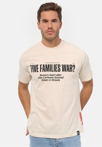 Camiseta de algodón color crema con texto negro destacado. Incluye detalles como una etiqueta roja en el dobladillo y una pequeña etiqueta de logo negra. Manga corta.