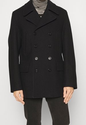 Homme portant un caban noir croisé avec de larges revers, un col roulé à rayures et un pantalon noir.