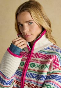 Strickpullover in Creme mit bunten geometrischen Mustern. Verfügt über einen hohen Kragen, einen pinken Reißverschluss und eine gerippte Textur an den Ärmeln.