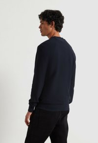 Navy pullover sweatshirt met geribbelde textuur, ronde halslijn en lange mouwen. Model draagt zwarte broek en staat met de rug naar de camera.