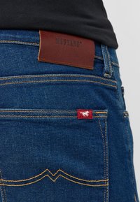Jeans dżinsowe w ciemnoniebieskim kolorze, z pomarańczowymi szwami, brązową skórzaną metką Mustang oraz małą czerwoną etykietą z emblematem konia.