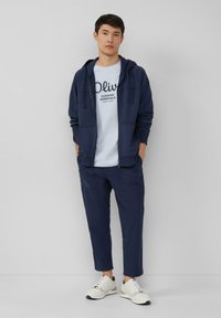 Marineblauwe zip-up hoodie over een lichtblauwe T-shirt met grafische tekst, gecombineerd met marineblauwe cropped broek en lichte sneakers. Glad fabric, casual pasvorm.