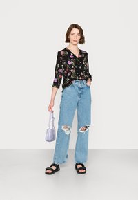 Blusa preta com padrão floral e mangas três quartos, jeans de perna larga rasgados em azul claro, sandálias pretas e uma pequena mala lavanda.