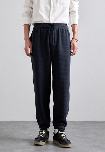 Pantaloni jogger de culoare bleumarin închis, confecționați dintr-un material texturat, cu talie elastică și șnur de ajustare. Asortați cu adidași negri, cu accente albe.