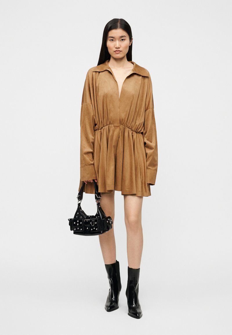 Braunes Wildleder-Kleid mit langen Ärmeln, tiefem V-Ausschnitt und elastischer Taille, kombiniert mit einer kleinen schwarzen Handtasche mit silbernen Beschlägen und Details.