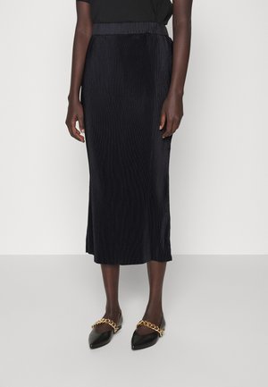 Femme portant une jupe midi plissée noire et des chaussures noires pointues avec de grands détails de chaîne dorée, debout devant un fond blanc.