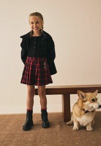 Černá košile na knoflíky, červená kostkovaná plisé sukně s kovovými sponami, černé kotníkové boty se zřasenými červenými akcenty, sedící corgi na dřevěné lavičce.