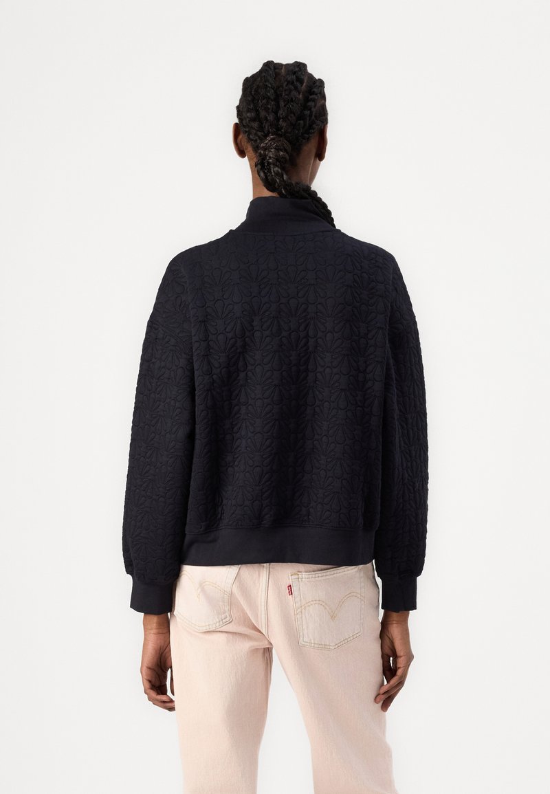 Sudadera negra acolchada con cuello y mangas de canalé, que presenta un patrón floral texturizado, llevada sobre unos vaqueros rosas claros.