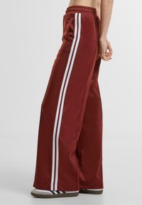 Maroon wide-leg pants with white side stripes, featuring an elastic waistband and smooth fabric texture. Accented with sporty stripes near the hem.
