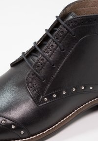 Chaussure habillée en cuir noir avec détails brogue, clous métalliques et lacets noirs sur une semelle marron cousue.