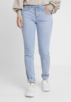 Vrouw draagt lichtblauwe opgerolde skinny jeans, beige trui en witte sneakers, staat met één hand in de zak.