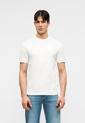 Άνδρας που στέκεται αντικρίζοντας μπροστά, φοράει απλό λευκό κοντομάνικο μπλουζάκι και μπλε τζιν, με φόντο φωτεινό γκρι.