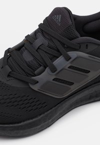Scarpa sportiva nera con tomaia in rete, lacci spessi, suola texturizzata e supporto in plastica nella zona mediana del piede con accenti a tre strisce laterali.