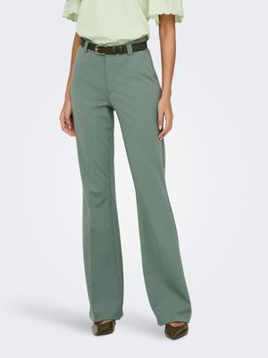 Frau trägt hoch taillierte, breitbeinige Hosen in Salbeigrün, hellgrüne Bluse, schwarzen Gürtel und olivfarbene spitze Schuhe, steht vor weißem Hintergrund.