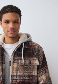 Chaqueta de cuadros marrón y crema con forro de sherpa, bolsillos delanteros y una sudadera con capucha gris debajo. Tejido texturizado con detalles de botones.