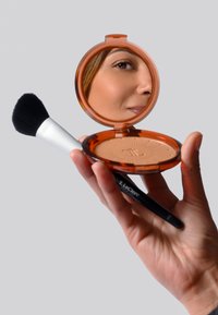 Bronzer-Compact mit einem orangefarbenen Etui, einem reflektierenden Spiegel und einem eingeprägten Design; daneben ein schwarzer Makeup-Pinsel mit einem weißen Griff.