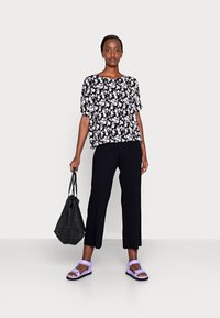 Top preto com padrão floral e mangas curtas, calças pretas caneladas com rachas laterais, sandálias lavanda e uma bolsa de mão preta texturizada.