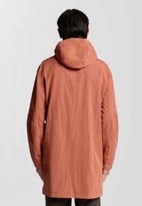 Longue veste légère de couleur corail atténuée avec une capuche, présentant une surface texturée et des manches amples. Design simple et minimaliste.