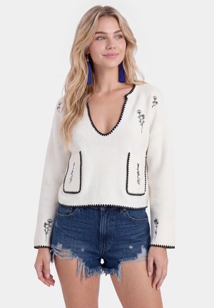 Ung kvinde med langt blondt hår iført hvid V-hals sweater med sort blomsterbroderi og slidte denimshorts.