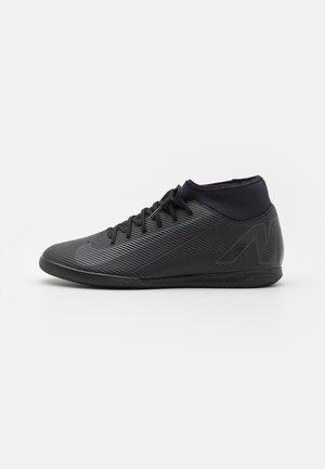 Chaussure de football en salle noire basse avec surface texturée, lacets et col rembourré, vue de côté gauche sur fond blanc.