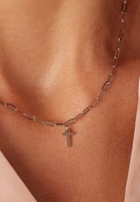 Zilveren ketting met langgerekte schakels en een klein kruisje als hanger. Vloeiende, reflecterende oppervlakte en een minimalistisch ontwerp.