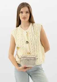 Metalická stříbrná crossbody taška s texturovaným povrchem, dvojitými přihrádkami a tenkým popruhem. Nosená s krémovým pleteným svetrem a světlými džíny.