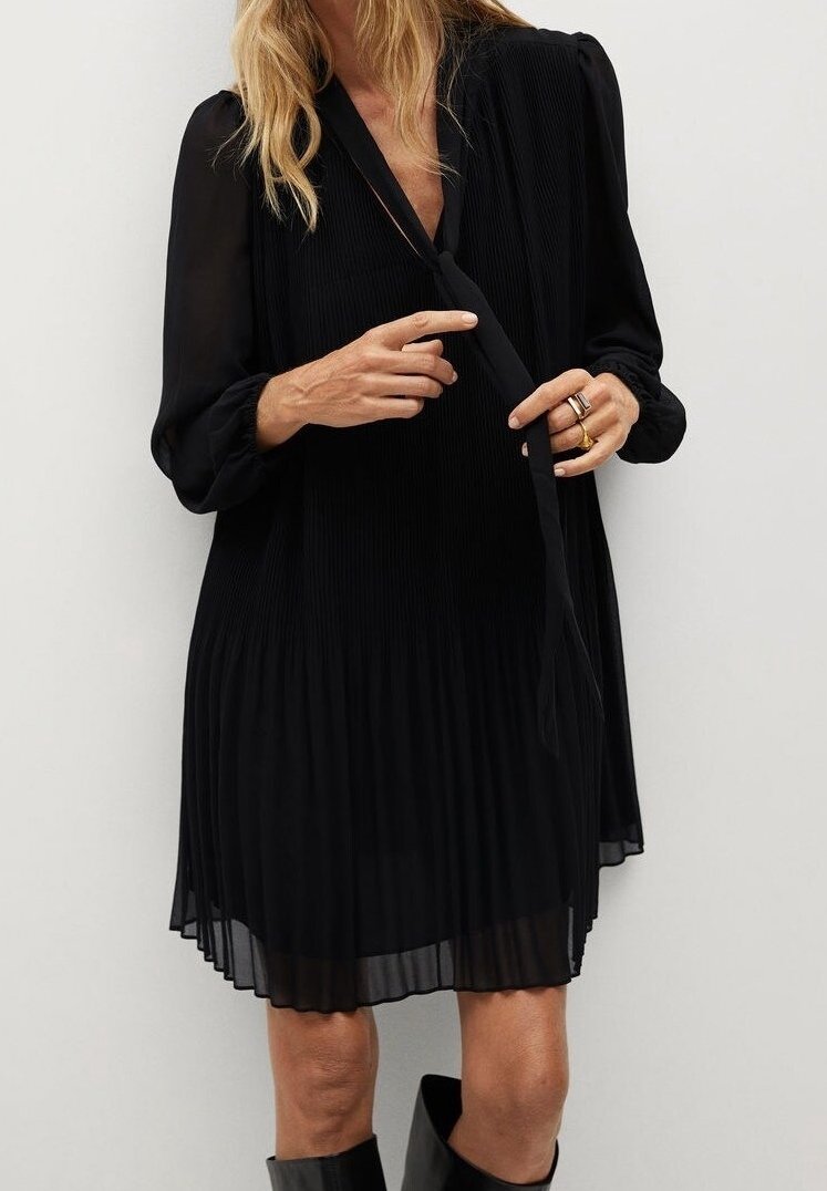 Vestido negro plisado con una tela suave y fluida, mangas largas y un escote en V profundo, con un detalle de lazo en la cintura.