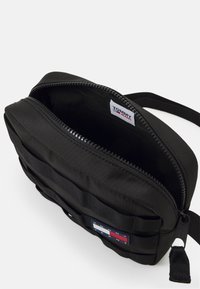 Borsa a tracolla nera in nylon con chiusura a zip, caratterizzata da un'esterno testurizzato, una tracolla regolabile e una patch logo sul fronte.