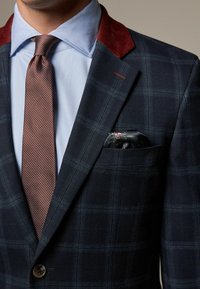 Marineprikket blazer med burgunderrød velourkrave, blå skjorte, tekstureret brun slips og mønstret lommehåndklæde i lommen.