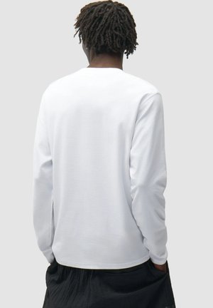 Persona con capelli scuri che indossa una maglietta bianca a maniche lunghe e pantaloni neri, in piedi con le mani in tasca, di spalle alla fotocamera.