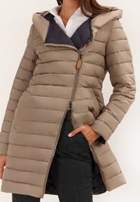 Žena v hnědé vyp padded dlouhé bundě s diagonálním zipem, ruka v kapse, pod bílou košilí a černobílými kostkovanými kalhotami.