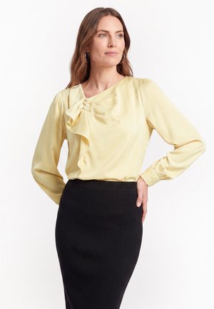 Femme portant un chemisier jaune pâle avec un détail en perles et une jupe noire cintrée, debout, une main sur la hanche, regardant vers la droite.
