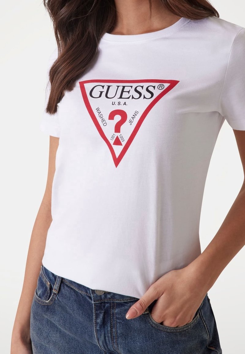 Camiseta de algodón blanca con un gráfico triangular rojo que presenta el texto "GUESS U.S.A." y un signo de interrogación. Combinada con unos jeans de mezclilla azul.