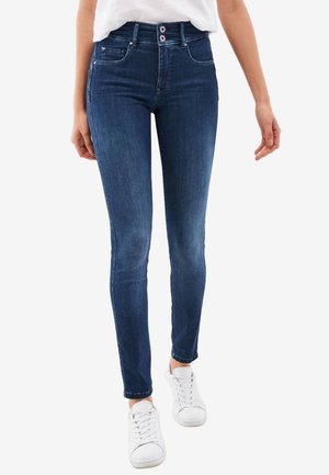 Woman wearing high-waist dark blue skinny jeans with two-button closure and white sneakers, standing against a white background.