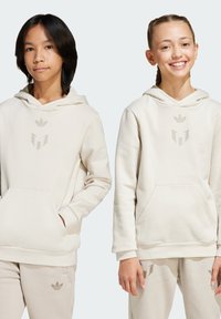 Les hoodies en coton beige sont dotés d'une poche avant, d'une capuche avec cordon de serrage et de deux logos sur la poitrine, portés avec un pantalon de jogging beige coordonné.