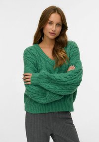 Pull en tricot torsadé vert avec un décolleté en V, coupe oversize et poignets côtelés. Porté sur un pantalon gris à rayures. La texture semble douce et épaisse.