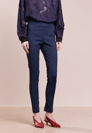 Mujer vestida con pantalones azul marino ajustados, una blusa estampada de color púrpura oscuro y zapatos slingback rojos de tacón bajo, de pie sobre un suelo beige.