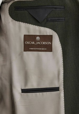 Intérieur de blazer vert avec une étiquette beige à couture marron. Comprend des accents noirs et une poche unique, avec un doublure beige lisse.