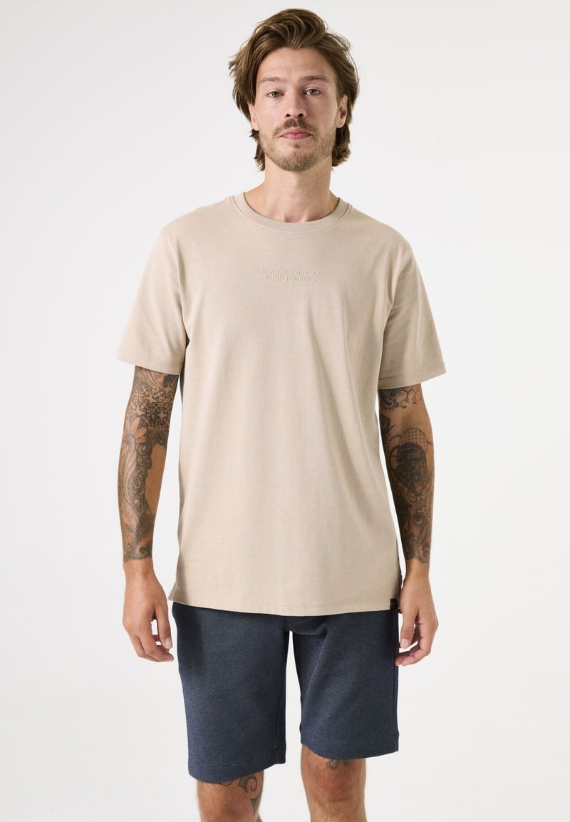 Beige katoenen t-shirt met korte mouwen, ronde halslijn en subtiele tekstdetails; gecombineerd met donkerblauwe shorts, relaxed fit.