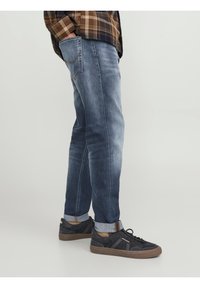 Man står med händerna i fickorna, iförd uppvikta, bleknade blå jeans, svarta sneakers med bruna sulor och en brun rutig skjorta.