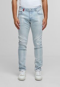 Vyras vilkintis šviesiai mėlynus suplėšytus džinsus, baltus sportinius batelius ir balta be rankovių marškinėlius, stovintis prieš paprastą pilką fono.